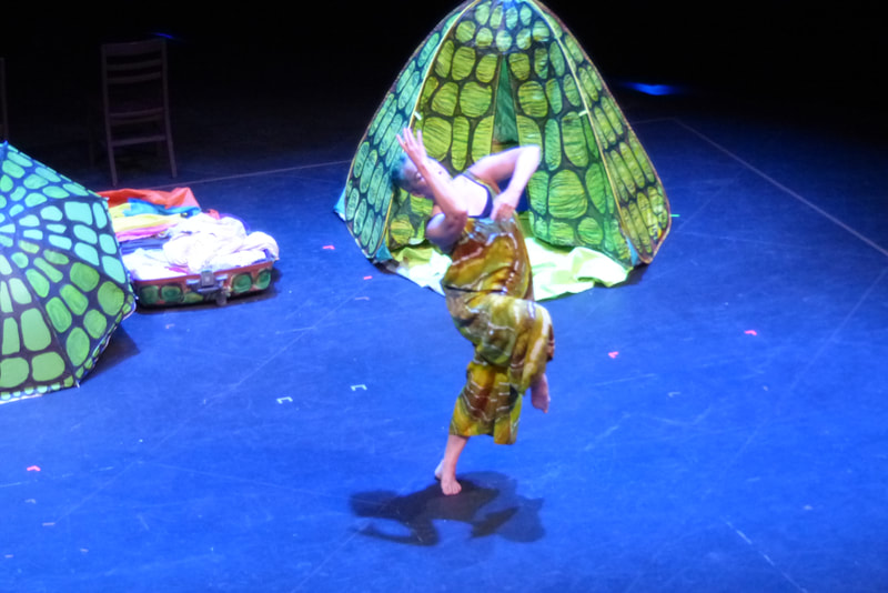 A dancer wearing brown green and white lifts one leg with the knee bent and leans it across her body her arms are gesturing back over her right shoulder and a pop up tent painted to look like a turtle shell is behind her. 