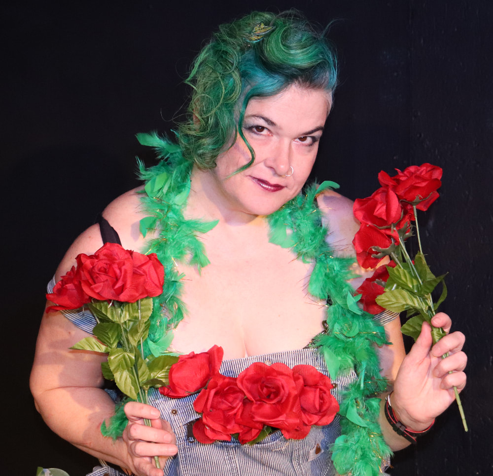 A white woman with green hair looks coyly into the camera. She is wearing overalls, a green feather boa,  and holding roses. 