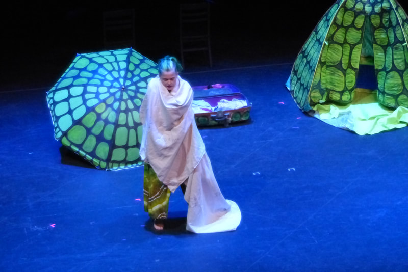 A dancer stands on stage, wrapped in a white cloth. An umbrella and pop up tent painted to look like turtle shells are behind her. 