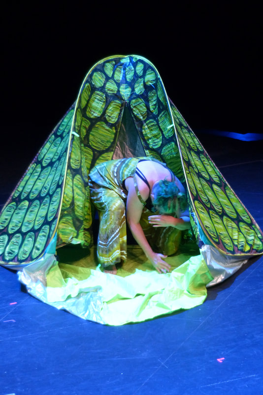 A green haired white woman bends over and gestures with her arms in the door of a small pop up tent painted to look like a turtle shell