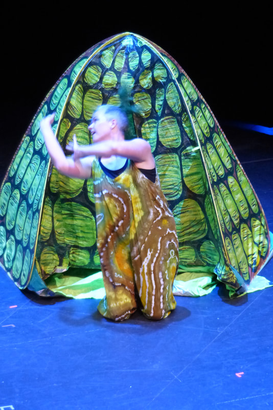 A dancer wearing a brown green and white jumpsuit gestures over her right shoulder in front of a pop up tent painted to look like a turtle shell.