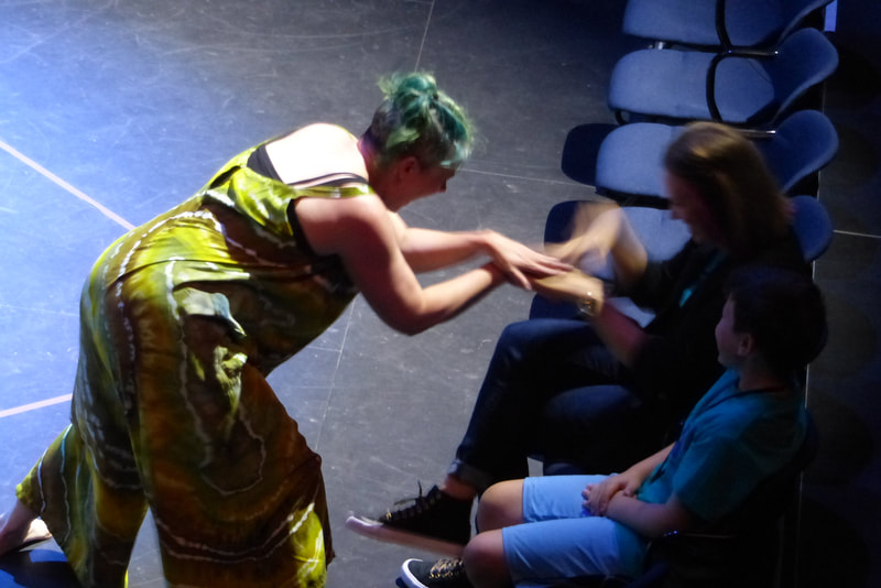 A dancers in a green brown and white tie dyed jumpsuit reaches toward and audience members, connecting their hands. 