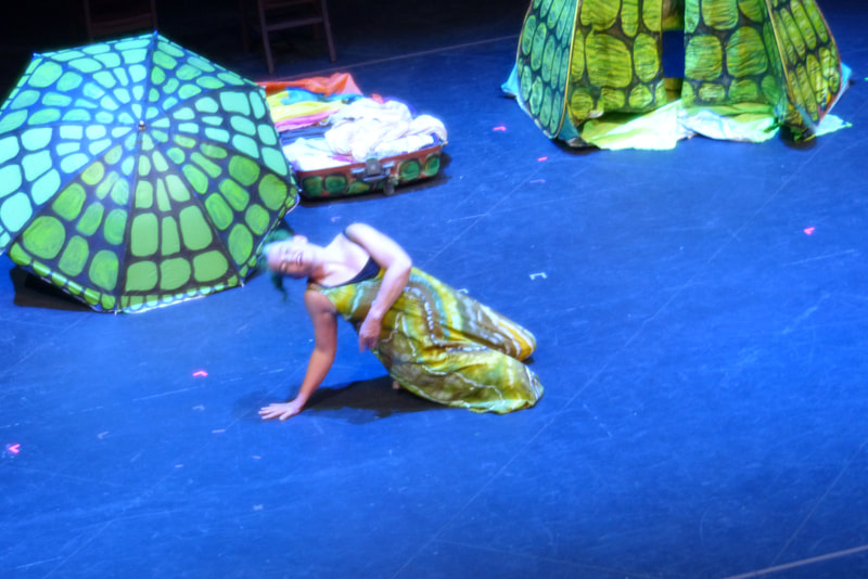 A dancer in a green brown and white tie dyed jumpsuit sits on her knees on the floor, leaning back onto one hand and wrapping her other arm around her midsection. A tent and umbrella painted to look like turtle shells are in the background. 